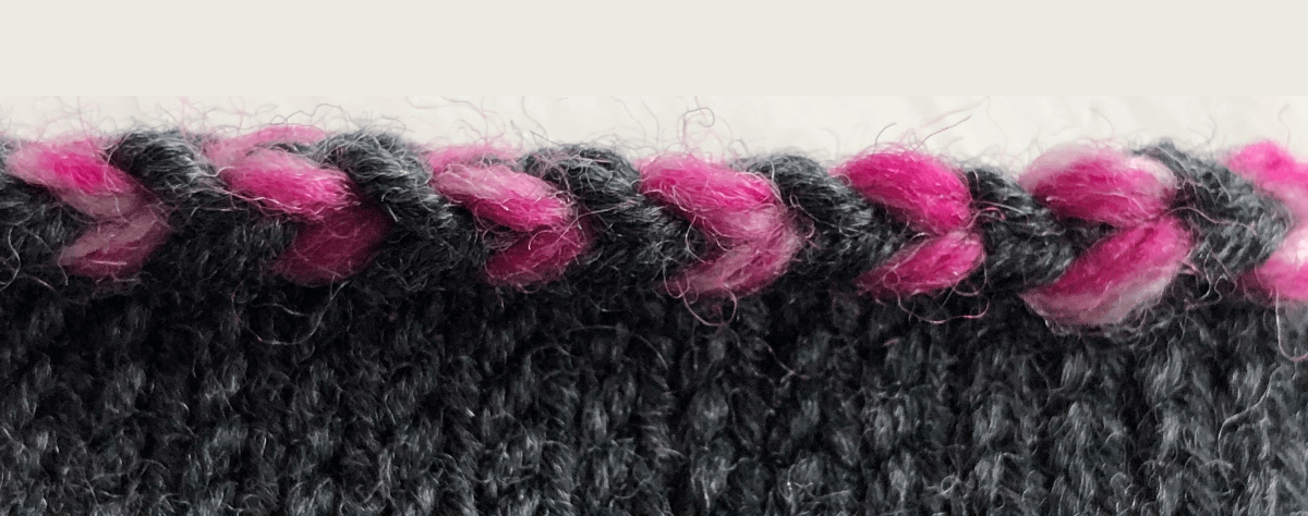 A close up of a the Latvian braid bind off. Stockinette stitch in dark grey with the bind off done in the dark grey and a variegated bright pink yarn.